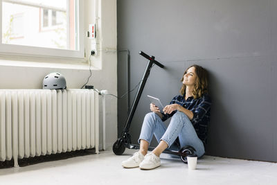Young woman with e-scooter sitting on floor, using digital tablet