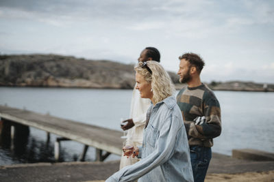 Side view of young woman enjoying drinks while walking with friends near lake