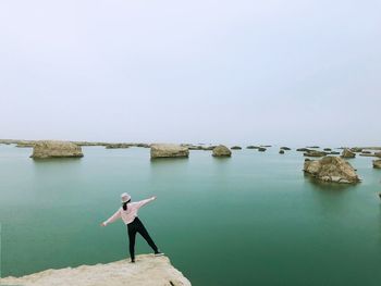 Woman with arms outstretched standing on rock at beach against clear sky