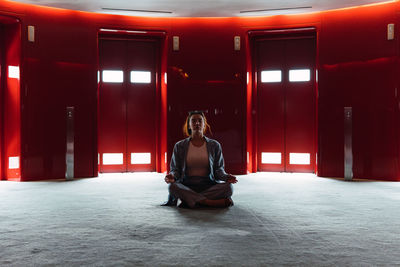 Full body of peaceful female sitting in lotus pose while meditating in spacious foyer with red elevators with glowing lights