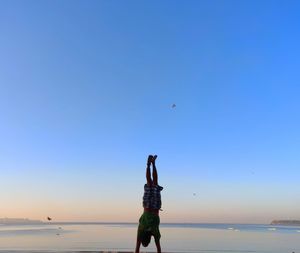 Full length of man practicing handstand at beach against clear sky during sunset