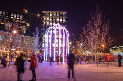 People on walkway against illuminated buildings in city at night