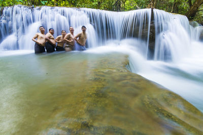 Cheerful shirtless friends standing at waterfall in forest