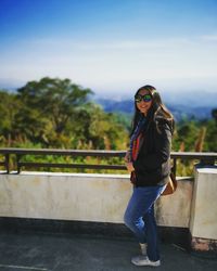 Portrait of young woman standing by railing against sky