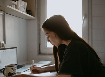 Side view of woman using laptop abd writing notes at home