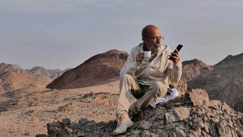 Senior man sitting on rock with smart phone and coffee 