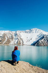 Rear view of young woman crouching by lake against mountain range