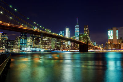 Illuminated bridge over river by buildings against sky at night