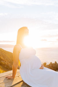 Pregnant woman sitting by sea against sky during sunset