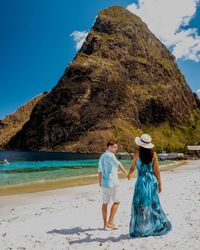 Rear view of couple holding hands while standing at beach against mountain