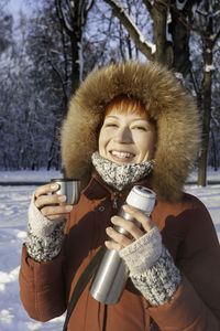 Smiling woman in red coat drinks hot tea from metal thermos. leisure activity in winter forest