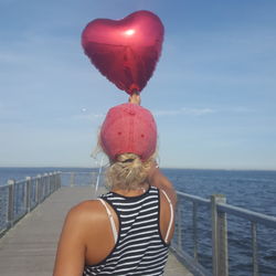 Rear view of woman holding red heart shape balloon on pier over sea against sky