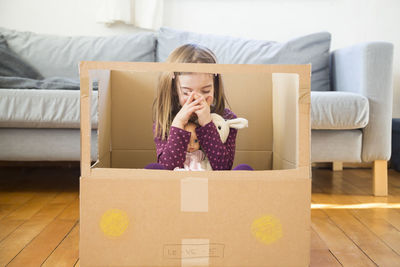 Girl with doll sitting in self-made cardboard car at home