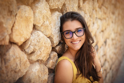 Young woman standing in front of a stone wall. wearing glasses smiling at the camera