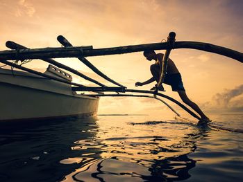 Shirtless man climbing outrigger on sea during sunset