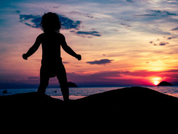 Silhouette small child standing by sea against sky during sunset