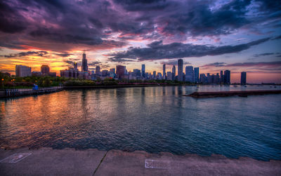 Sea by modern buildings against sky at sunset