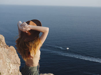 Rear view of woman with hands behind head wearing hat while standing on rock formation against sea