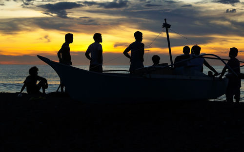 Silhouette people with boat at beach against sky during sunset