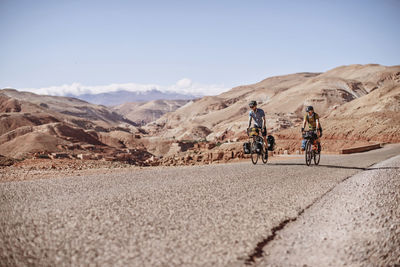 Two bike packers ride along a road with desert mountains behind them