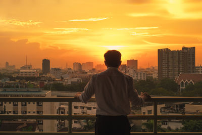 Rear view of man standing by buildings against sky during sunset