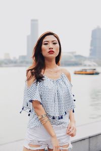 Portrait of young woman standing on footpath against river in city