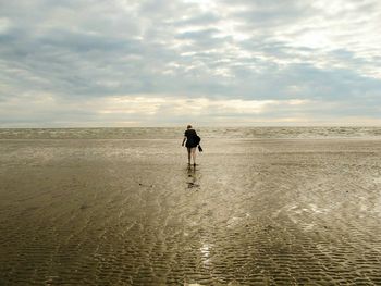 Rear view of woman walking on shore at beach against cloudy sky
