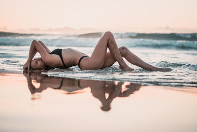 Side view portrait of seductive woman lying on shore at beach against sky during sunset