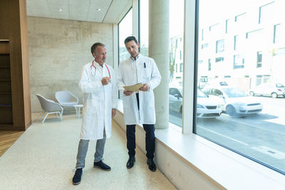 Doctors in lab coats discussing over medical record standing by window at hospital