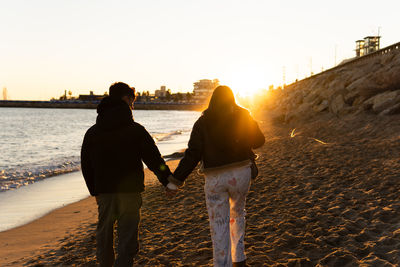 A young couple is walking along the beach holding hands as the sun sets.