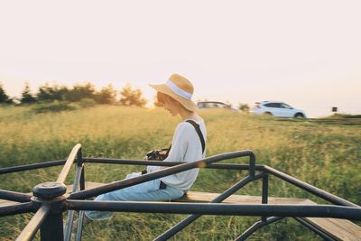 Smiling young woman sitting on wooden plank over grassy field against clear sky