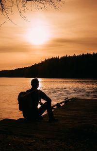 Rear view of silhouette man sitting by lake against sky during sunset