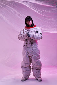 Full body asian woman in spacesuit adjusting gloves while standing against futuristic background under pink neon light