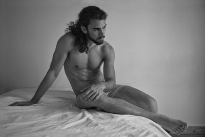  shirtless young man looking away while sitting on bed at home