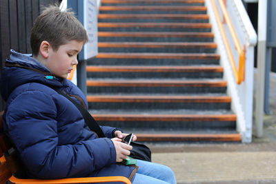 Side view of boy using mobile while sitting on bench at park