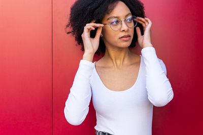 Serious attractive african american female with black hair in white clothes putting on stylish eyeglasses while standing on red background