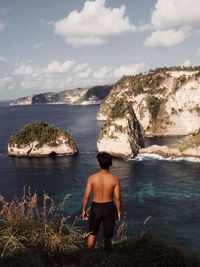 Rear view of shirtless young man looking at sea while standing against sky