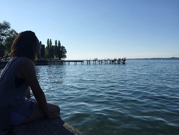 Rear view of woman sitting by sea against clear sky