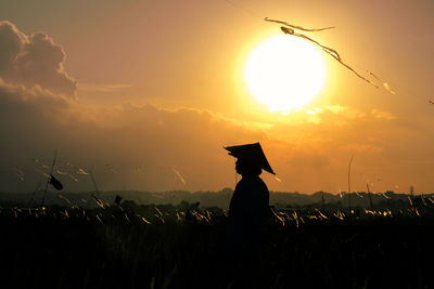 Silhouette woman with arms outstretched standing on field against orange sky during sunset