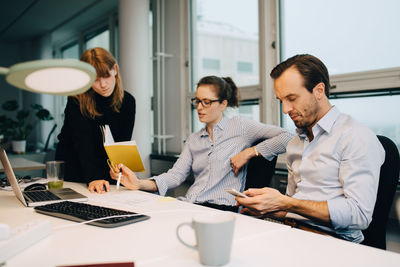 Businessman using smart phone while female colleagues discussing over document at desk in creative office