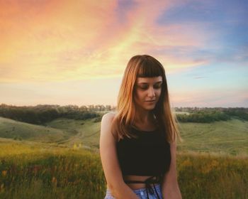 Beautiful young woman standing on field against sky during sunset