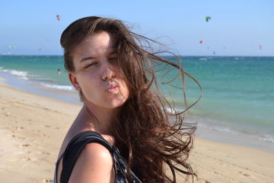 Beautiful young woman with tousled hair puckering lips at beach against sky