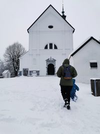 Rear view of person walking on snow covered field against building