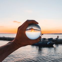 Close-up of hand holding crystal ball against sea during sunset