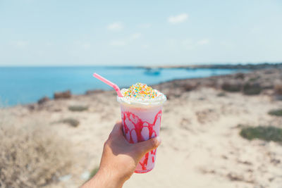 Cropped hand holding ice cream in glass at beach against sky