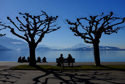 Silhouette people sitting by lake against clear blue sky