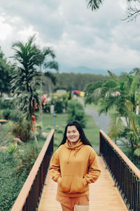 Portrait of young woman standing on footbridge against sky
