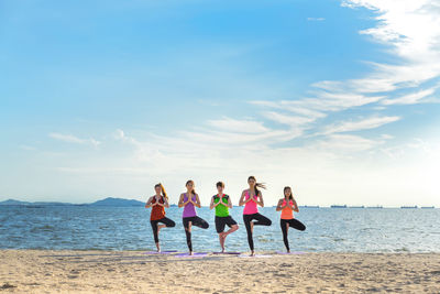 Young friends practicing yoga in tree pose on shore at beach during sunny day