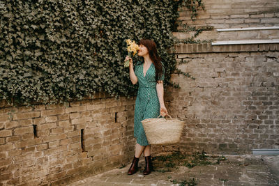 Full length of woman smelling flowers while standing against creeper wall