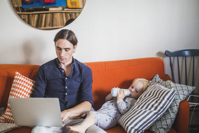 Stay at home father using laptop while sitting by toddler son drinking milk from bottle on sofa in apartment